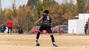 A soccer player wearing number 6 dribbling during a daytime match on a dry, grassy field.