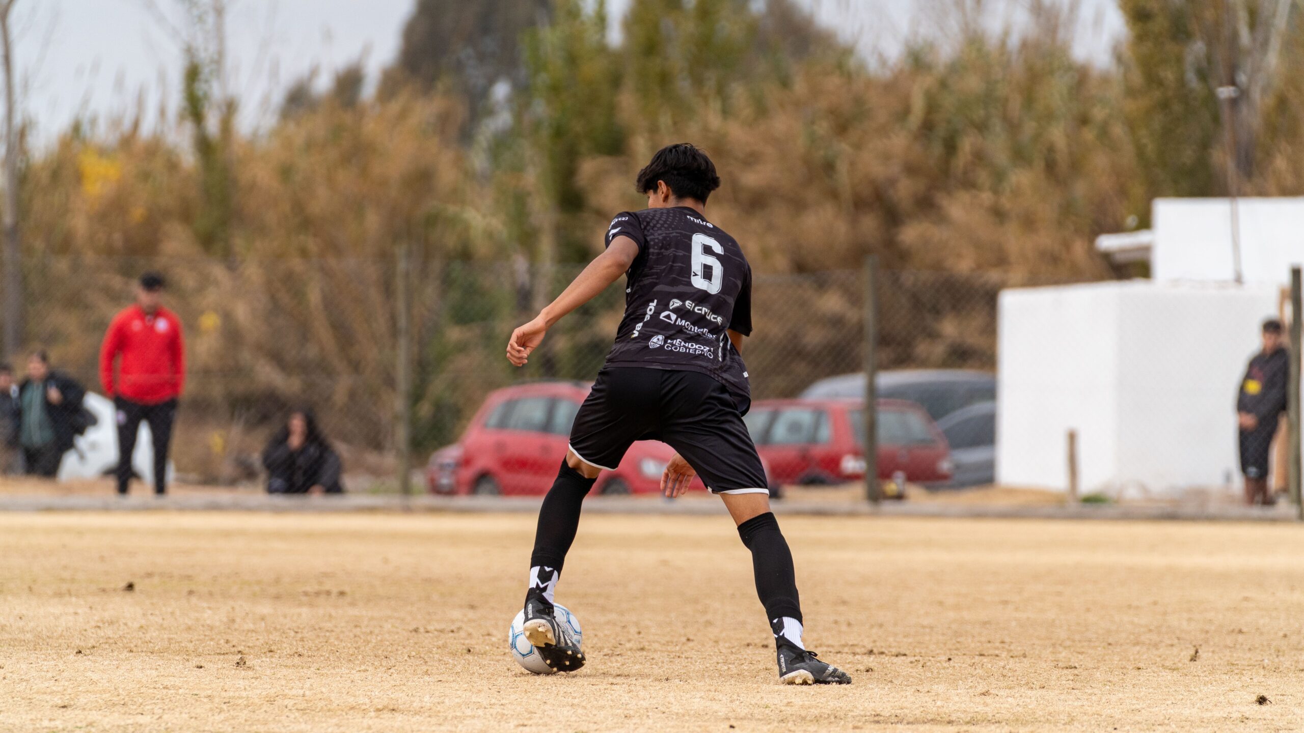 A soccer player wearing number 6 dribbling during a daytime match on a dry, grassy field.